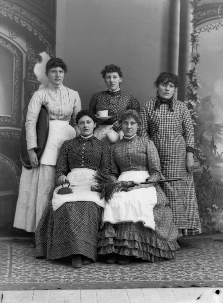 Five women pose for a group portrait in front of a painted backdrop with various household articles such as an iron, feather duster, platter, cup, pitcher, and fork. They are wearing long dresses and aprons.