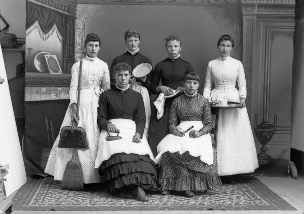 A studio portrait of six women posing in front of a painted backdrop. They are holding household items including an iron, knife, potato, dustpan, broom, pie dish, towel, plate, platter, cup, and saucer. They are wearing long dresses, and two wear aprons.