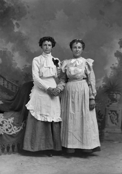 Two women stand for a studio portrait in front of a painted backdrop. They are wearing aprons over their long dresses and are holding hands.