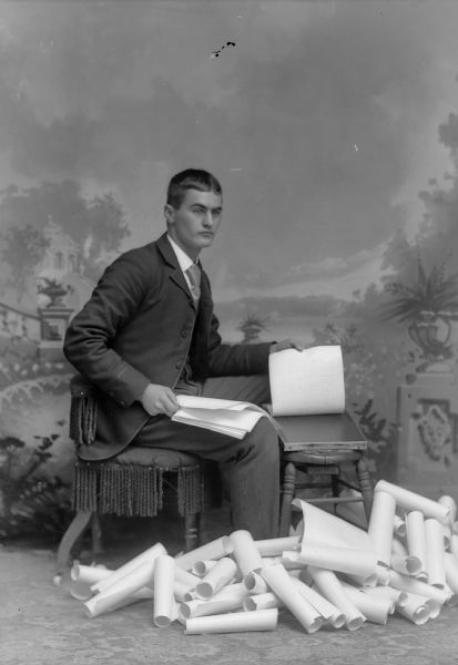 A studio portrait of a seated man on a chair wearing a suit jacket, vest, and trousers. He is demonstrating gelatin duplicating equipment on paper in front of a painted backdrop.