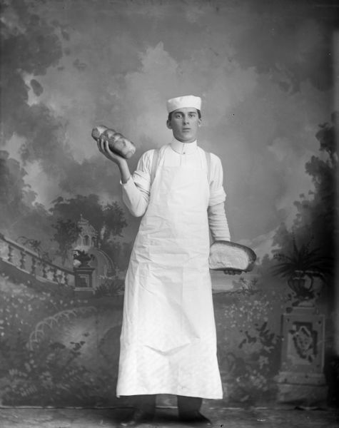 A baker wearing a long apron holds two loaves of bread while posing for a studio portrait in front of a painted backdrop.