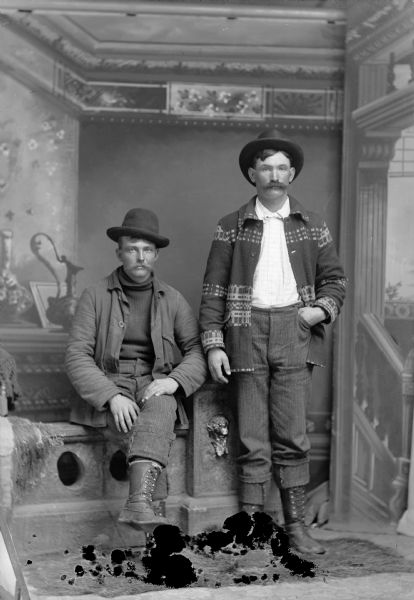 Two workmen, one seated on a low stone wall, and one standing, pose for a studio portrait in front of a painted backdrop. The men may be workmen or lumberjacks, as they are dressed in wool clothes, hats, and heavy boots.