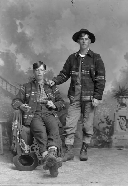 A studio portrait of two men, possibly lumberjacks, wearing wool jackets and heavy boots in front of a painted backdrop. The man sitting is smoking a cigar.