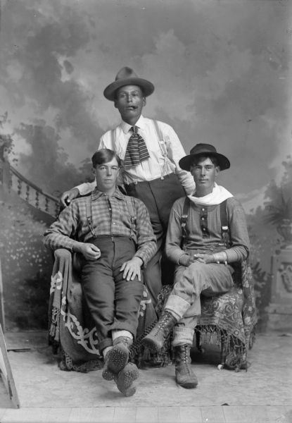 A studio portrait of three men in front of a painted backdrop. Two sitting men, possibly lumberjacks, wear wool clothes with suspenders and heavy boots, while another man stands behind them.