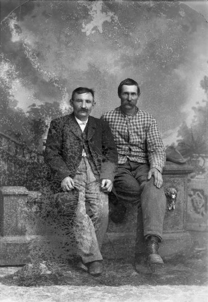 Two men sit on a low stone wall and pose for a studio portrait in front of a painted backdrop.