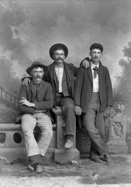 A studio portrait of three men posing near a stone wall in front of a painted backdrop. They are all wearing hats, suit jackets, and trousers.