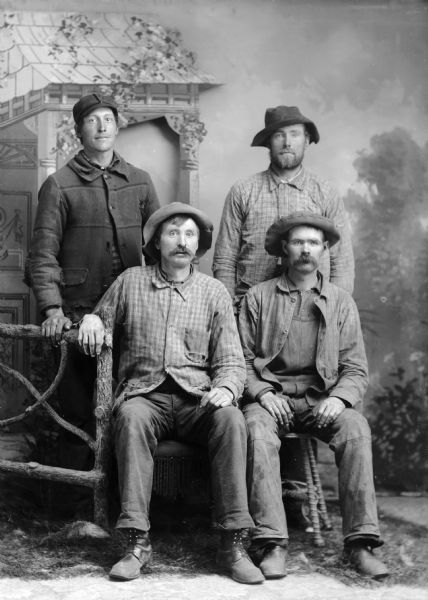 Four workmen, two sitting and two standing, pose for a studio portrait in front of a painted backdrop. They are all wearing hats, and there is a wood rustic fence on the left.