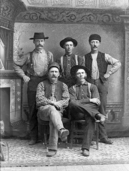 Five men, two seated and three standing, pose for a studio portrait in front of a painted backdrop.