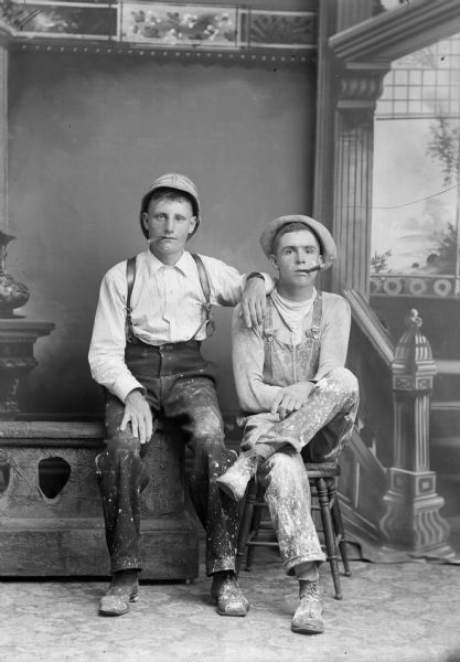 A studio portrait of two seated men, one on a chair and the other on a low stone wall, smoking cigars. They are wearing hats and paint-splattered clothes in front of a painted backdrop.