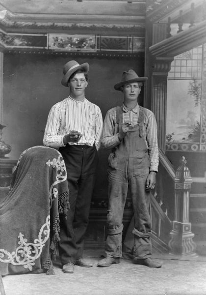Studio portrait of Jule (in overalls) and Joe Hubert. Both men are holding cigars and are wearing tilted hats in front of a painted backdrop.