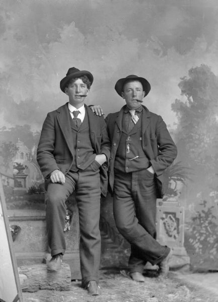 A studio portrait of two well-dressed men wearing hats, suit jackets, vests with watch fob, neckties, and trousers. They are smoking cigars in front of a painted backdrop.