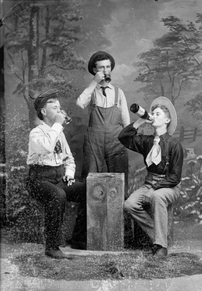 A studio portrait of three men sitting and standing around a crate in front of a painted backdrop and drinking. All three men are wearing hats and neckties. The man standing is wearing overalls.