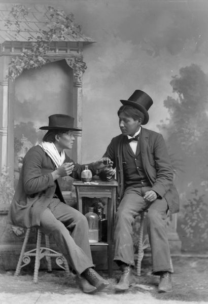 A studio portrait of two men, both wearing hats, seated at a small table, drinking and smoking in front of a painted backdrop. They are identified as Henry Badsoldier Stacy (MonNawPaeSheSheckKaw), left, and Will Stohegah Carriman (WonkShiekStoHeKah).