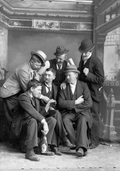 A studio photograph of six, well-dressed men gathered around two beer bottles, intently examining one of the bottles in front of a painted backdrop. Four of the men are wearing hats, and most wear suit jackets, vests, neckties, and trousers.
