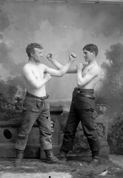 Two young men stand for a studio portrait in front of a painted backdrop. The men are shirtless, and wear trousers and boots. They are posing as if they were about to box.