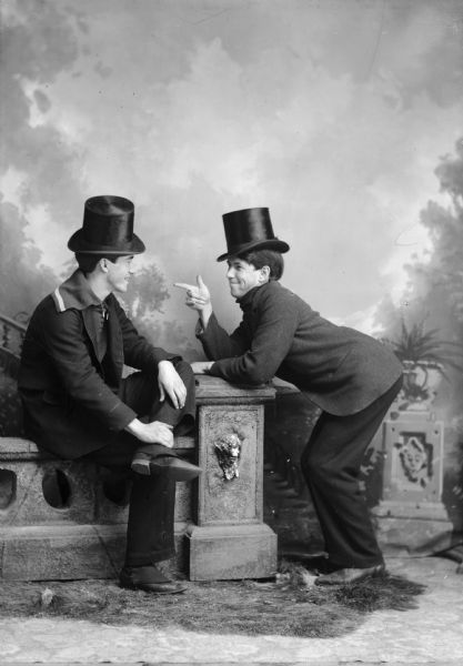 Studio portrait of two nattily-dressed men wearing top hats. One man is pointing at the other man. They are posing for the camera on a low stone wall in front of a painted backdrop.