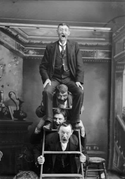 Four men use a ladder to pose on top of each other. There is a painted backdrop behind them. They are wearing suit jackets, vests with watch fobs, and trousers.