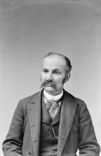 A well-dressed Charles J. Van Schaick poses for a self-portrait in his studio. He is wearing a suit jacket, vest with watch fob, and a necktie, and has a moustache and sideburns.