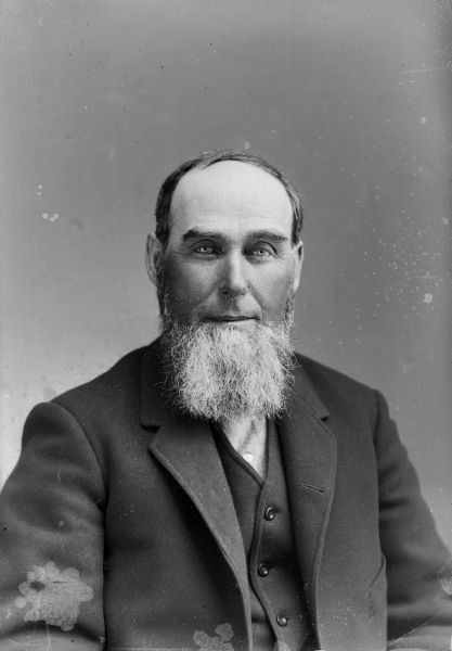 A bearded man with a suit jacket and vest sits for a studio portrait.