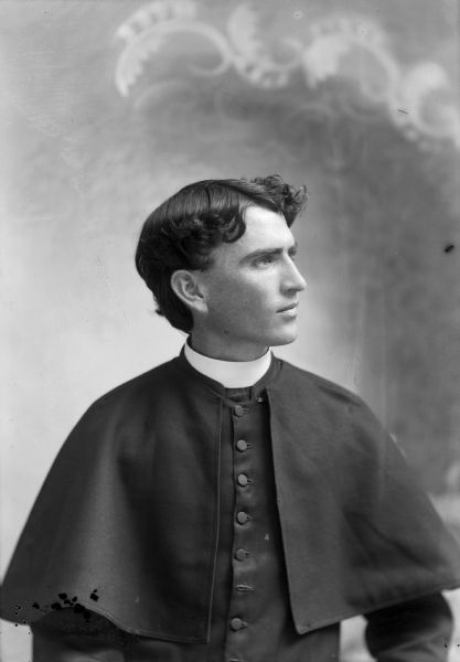 A studio portrait of a young man wearing a clergy robe, identified as Reverend Father P.F. Garrity. He is posing in front of a painted backdrop.