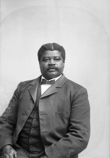 A studio portrait of a well-dressed, African-American man, identified as H.F. "Frank" Lyons. He is wearing a suit jacket, vest, and necktie. Lyons was the Water Street barber in Black River Falls.