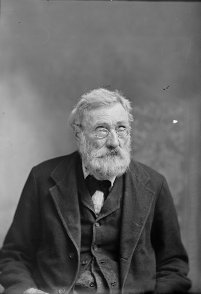 An elderly man sits for a studio portrait. He wears wire-rimmed eyeglasses, wears a suit jacket, vest, and necktie and has a beard and moustache.