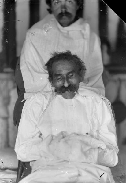 A portrait of a seated elderly man dressed in a white gown. Probably a corpse, the body is held in position by another man.