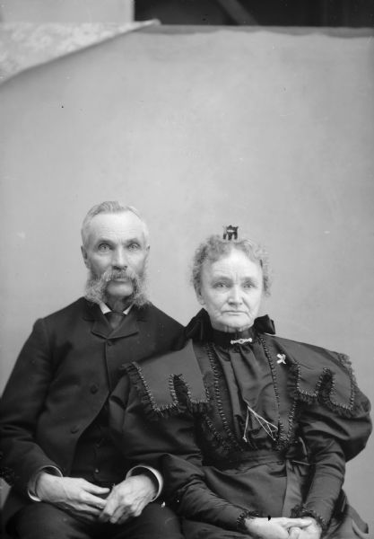 An elderly man and woman pose while sitting for a studio portrait in front of a backdrop. The man has muttonchops and is wearing a suit jacket and necktie. The woman has a hair comb or ribbon in her hair and wears a dress with an elaborately decorated collar and puffed sleeves.
