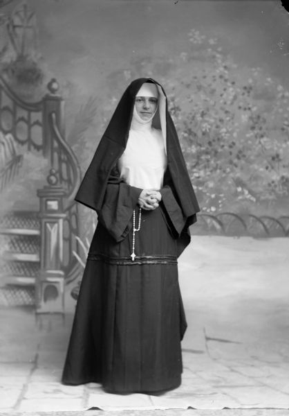 A nun folds her hands at her waist and stands for a studio portrait in front of a painted backdrop.