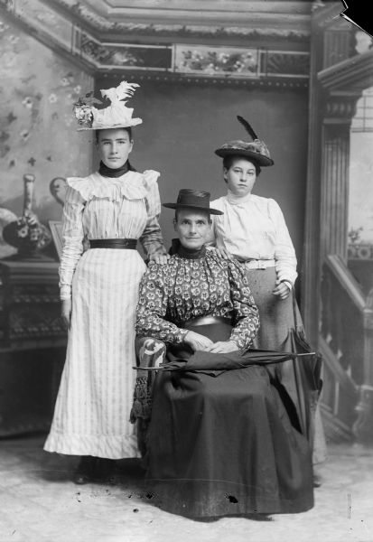 Two young women stand on each side of an older woman who sits with an umbrella resting on her lap. All three women wear hats as they pose for a studio portrait in front of a painted backdrop.