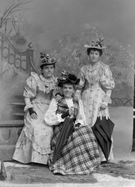 A studio portrait in front of a painted backdrop of three women wearing elaborately designed hats. Two of the women hold umbrellas.