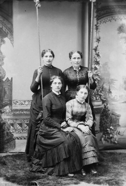 A studio portrait of four women in front of a painted backdrop. Two women sit on a swing while the other two hold the ropes of the swing.