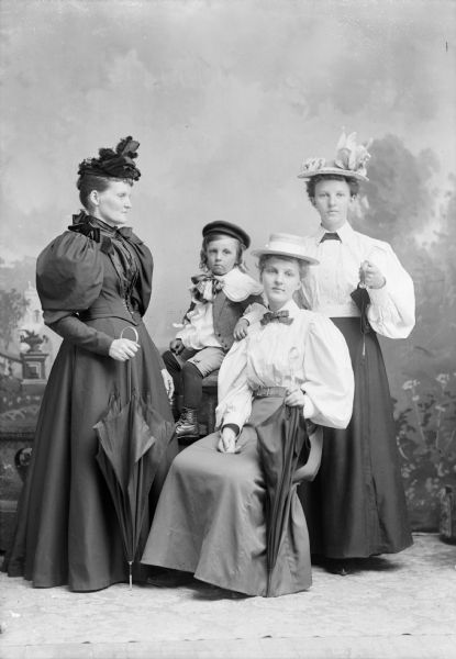 Three women and a child pose for a studio portrait in front of a painted backdrop. All of the women are holding umbrellas and are wearing hats. The child, wearing a hat, sits atop a table in the center.