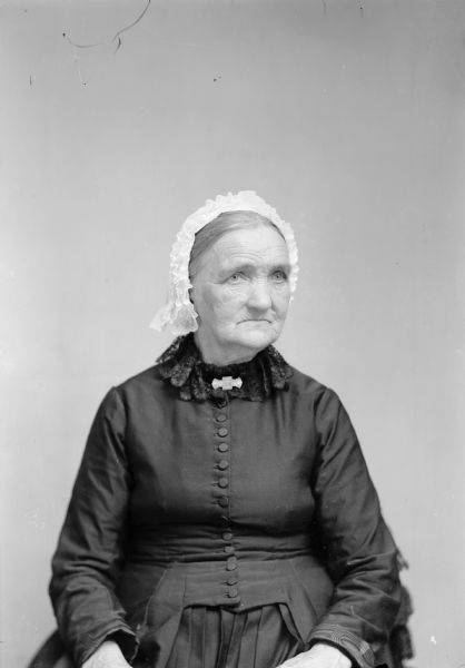 A seated elderly woman wearing a white, lace cap poses for a studio portrait.