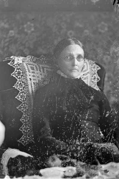 A studio portrait of an elderly woman sitting in a stuffed chair and wearing eyeglasses.
