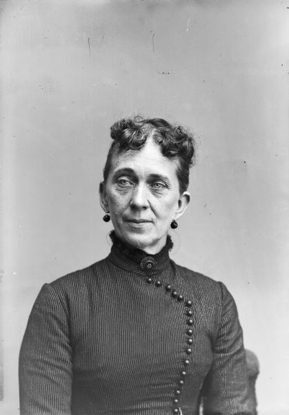 A seated woman wearing a black dress with buttons up the front, a brooch, and earrings, poses for a studio portrait.