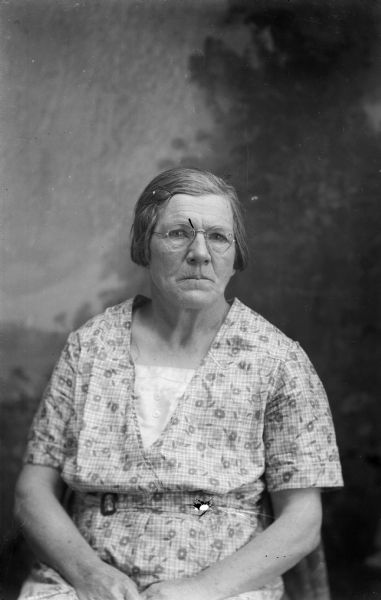 A studio portrait of a seated elderly woman wearing a casual dress in front of a painted backdrop.