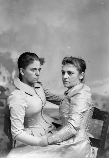 Two well-dressed women face each other and pose for a studio portrait in front of a painted backdrop.