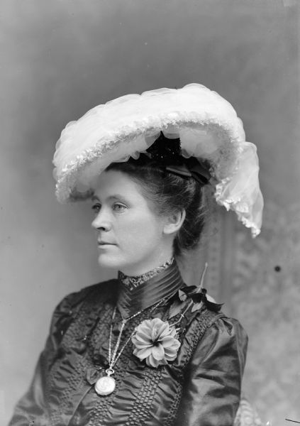 A well-dressed woman poses for a portrait. She wears a large white hat, a locket, and flower corsage.