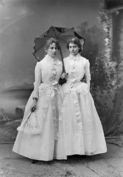 Two women in long white dresses stand for a studio portrait in front of a painted backdrop. One woman holds a parasol while the other holds a hand fan.
