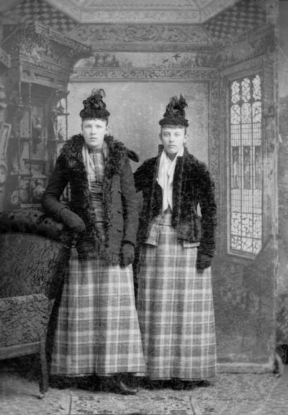 Two women, similarly dressed with feathered hats, black jackets, and plaid skirts, stand for a studio portrait in front of a painted backdrop.