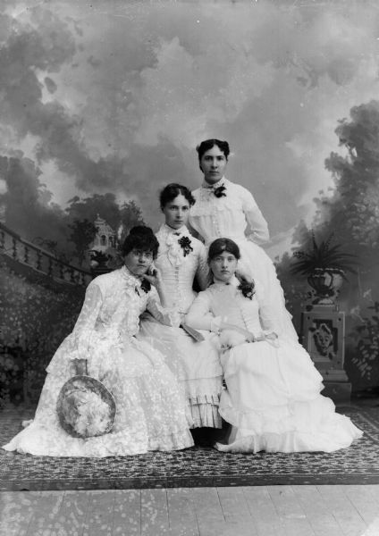 A studio portrait of four women in front of a painted backdrop. All of the women wear white dresses with pinned corsages on their shoulder.