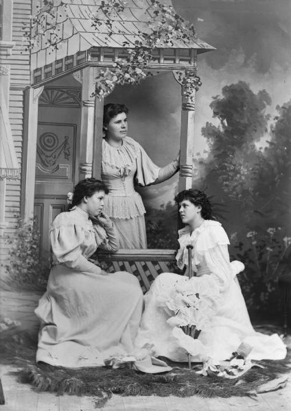 Studio portrait of three young women in dresses posing around a prop of a covered entrance to a house in front of a painted backdrop.
