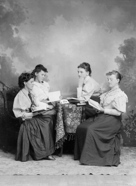 Four young women sitting around a table and reading books while posing for a studio portrait in front of a painted backdrop. They are wearing white blouses and long, black skirts.