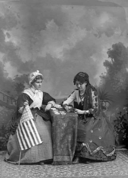 A studio portrait of two women sitting at a table in front of a painted backdrop. One of the women is dressed as a fortune teller and holds round cards. The other woman is wearing a bonnet and holding an American flag. It appears the woman is having her fortune told.