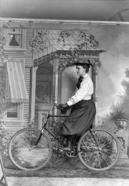 A studio portrait of a woman riding a men's bicycle in front of a painted backdrop of a house. She wears a skirt, riding gloves and a hat.