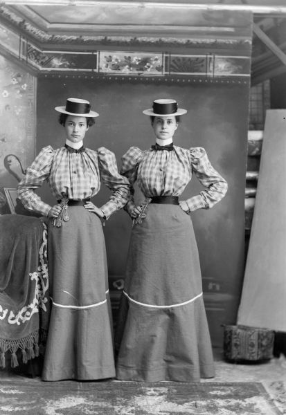 Full-length portrait of two women standing for a studio portrait in front of a painted backdrop. The women are dressed identically and are wearing skirts, belts, shirts, and hats.