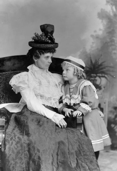 A studio portrait of a seated woman wearing a hat and a young girl leaning towards her in front of a painted backdrop.