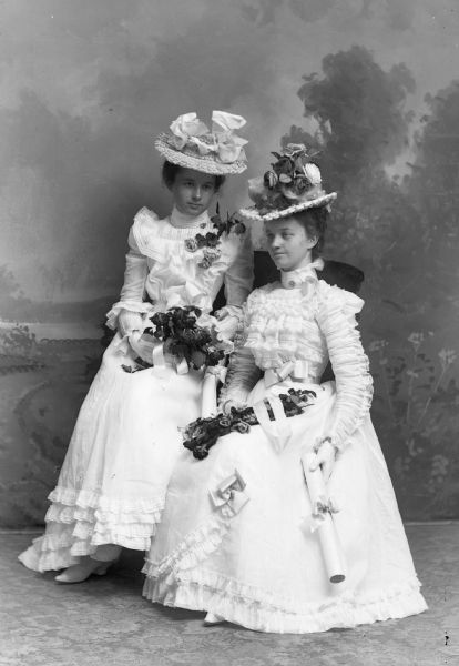 A studio portrait of two women wearing white dresses and elaborate hats in front of a painted backdrop. Both are seated on the same chair holding flowers, and each is holding a rolled piece of paper, perhaps a diploma.