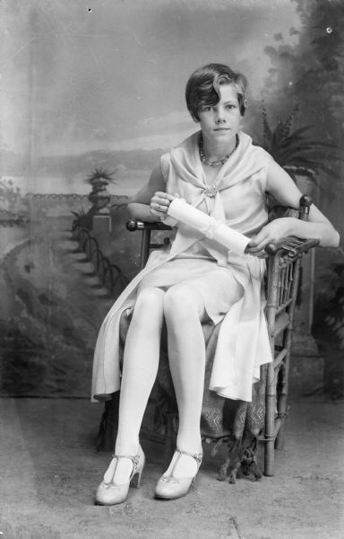A young woman with short hair sits in a chair and poses for a studio portrait in front of a painted backdrop. She holds a rolled piece of paper, perhaps a diploma.